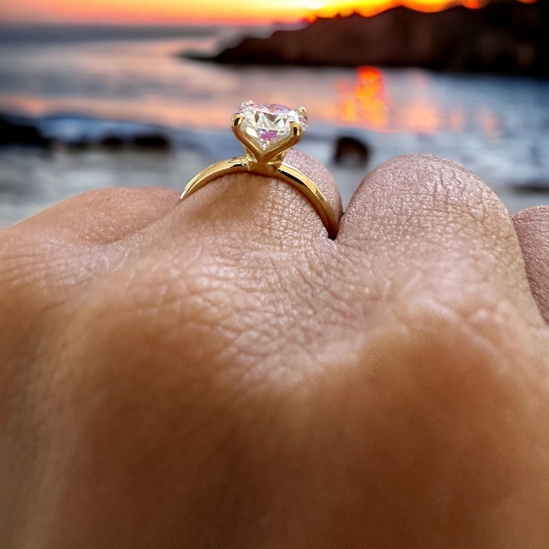 May include: A gold engagement ring with a large, clear, heart-shaped gemstone. The ring is set against a blurred background of a sunset over the ocean. The ring is on a finger.