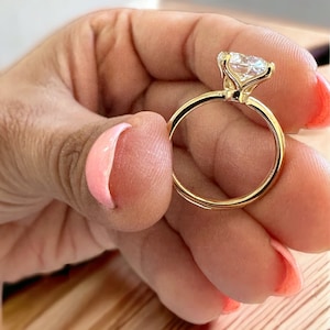 May include: A gold ring with a large, clear gemstone. The ring's band is a thin, polished gold, and the gemstone is held in place by delicate prongs. The ring is held between fingers with pink nail polish.