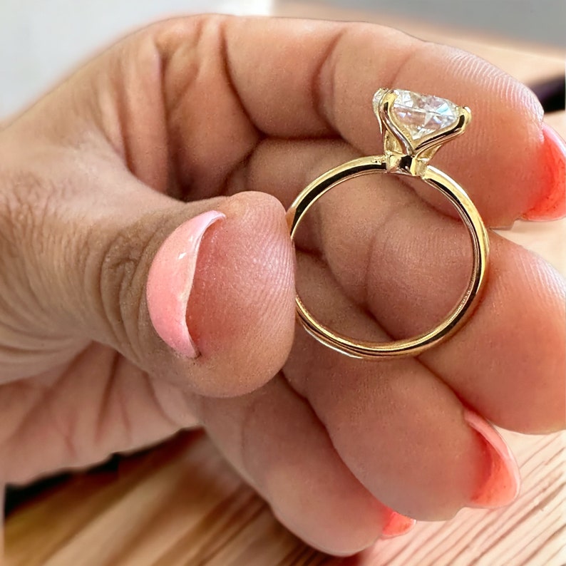 May include: A gold ring with a large, clear gemstone. The ring's band is a thin, polished gold, and the gemstone is held in place by delicate prongs. The ring is held between fingers with pink nail polish.