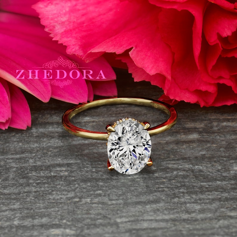 May include: A gold engagement ring with a large oval-shaped diamond. The ring is set on a gray wooden surface with pink flowers in the background.