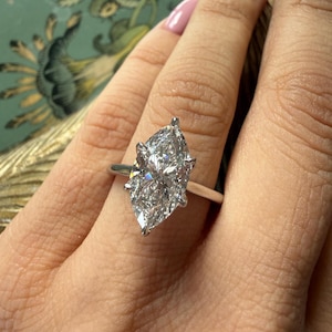 Pu&ograve; includere: Anello di fidanzamento con diamante taglio marquise e fascia argentata. Il diamante &egrave; il punto focale, che brilla intensamente. L'anello &egrave; su un dito con smalto rosa chiaro.