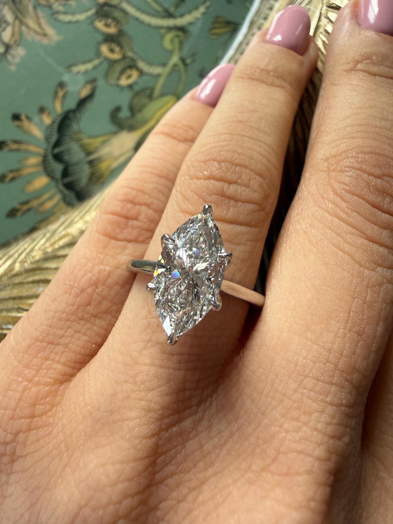 Pu&ograve; includere: Anello di fidanzamento con diamante taglio marquise e fascia argentata. Il diamante &egrave; il punto focale, che brilla intensamente. L'anello &egrave; su un dito con smalto rosa chiaro.
