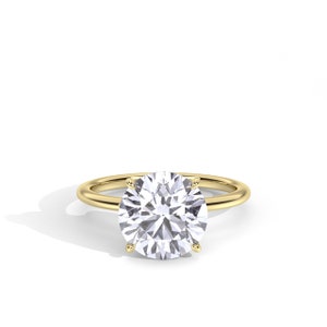 May include: A gold ring with a large, round, clear gemstone. The gemstone is held in place by four prongs. The band of the ring is a simple, polished gold. The ring is set against a white background.