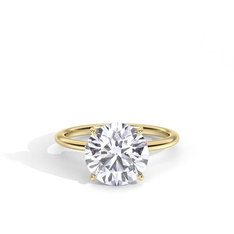 May include: A gold ring with a large, round, clear gemstone. The gemstone is held in place by four prongs. The band of the ring is a simple, polished gold. The ring is set against a white background.