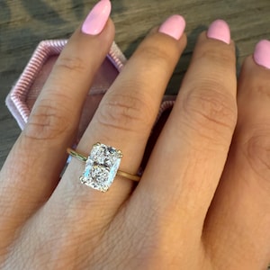 Puede incluir: Un anillo de oro con una gran gema clara rectangular. El anillo está en un dedo con esmalte de uñas rosa. El anillo está engastado en una banda de oro y se muestra en un joyero rosa.