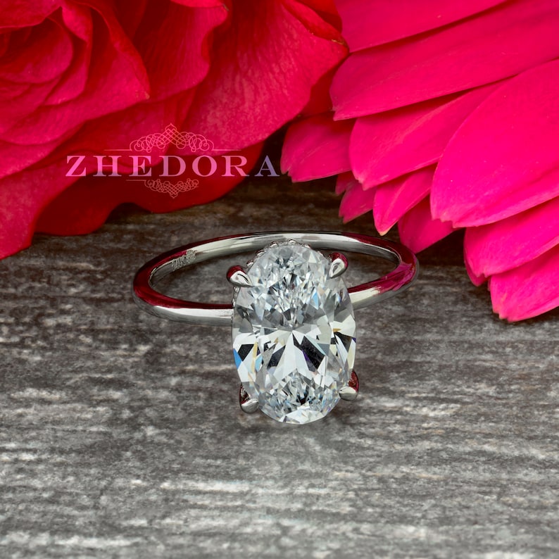 Puede incluir: Un anillo de plata con un solitario de diamante de forma ovalada. El anillo est&aacute; colocado sobre un fondo de p&eacute;talos de rosa rosados.