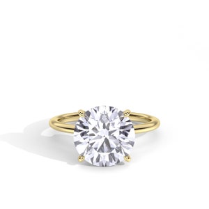 May include: A gold ring with a large, round, clear gemstone. The gemstone is held in place by four prongs. The band of the ring is a simple, thin gold band. The ring is set against a white background.