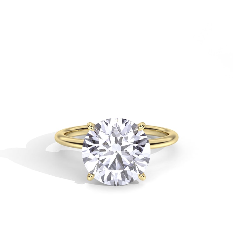 May include: A gold ring with a large, round, clear gemstone. The gemstone is held in place by four prongs. The band of the ring is a simple, thin gold band. The ring is set against a white background.
