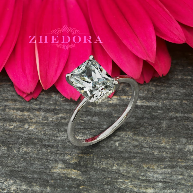 May include: A silver ring with a large, clear, square-shaped diamond. The diamond is set in a prong setting and has a halo of smaller diamonds around it.