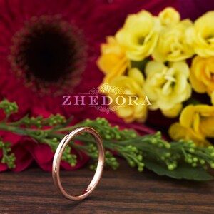 May include: A thin rose gold band ring, lying on a wooden surface with a blurred background of red and yellow flowers.