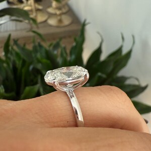 May include: A close-up of a silver ring featuring a large, oval-cut diamond. The diamond is set in a prong setting, and the band is smooth and polished. The ring is on a finger, with a blurred background of greenery.