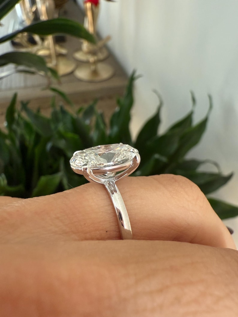 May include: A close-up of a silver ring featuring a large, oval-cut diamond. The diamond is set in a prong setting, and the band is smooth and polished. The ring is on a finger, with a blurred background of greenery.