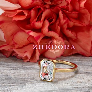 May include: A gold ring with a large, clear, rectangular-shaped gemstone. The ring is set against a background of red carnations and a wooden surface. The text "ZHEDORA" is visible in the image.