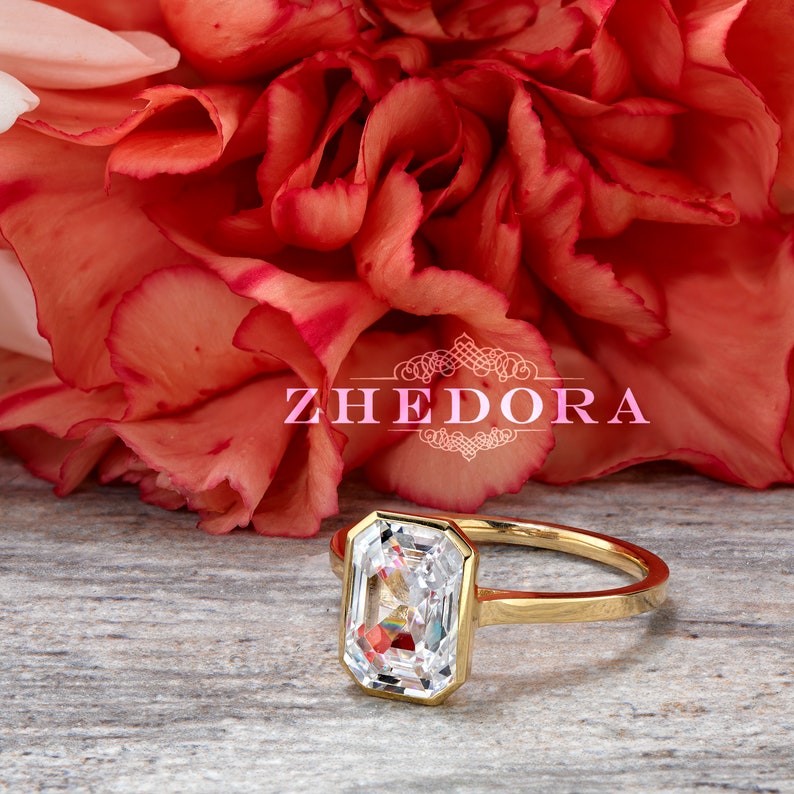 May include: A gold ring with a large, clear, rectangular-shaped gemstone. The ring is set against a background of red carnations and a wooden surface. The text "ZHEDORA" is visible in the image.