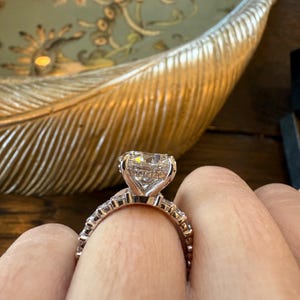 K&ouml;nnte beinhalten: Ein ros&eacute;goldener Verlobungsring mit einem gro&szlig;en, herzf&ouml;rmigen Diamanten als Mittelstein. Das Band ist mit kleineren Diamanten besetzt. Der Ring wird an einem Finger gezeigt, mit einem verschwommenen Hintergrund einer dekorativen Schale.