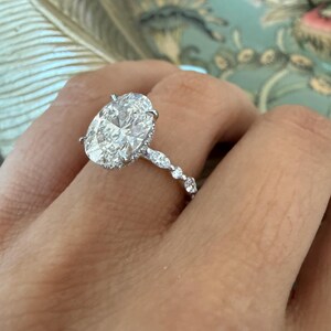 May include: A sparkling diamond ring with an oval-cut center stone. The ring features a delicate band with smaller accent diamonds. The setting is a bright, polished metal, likely platinum or white gold, and the ring is shown on a hand.