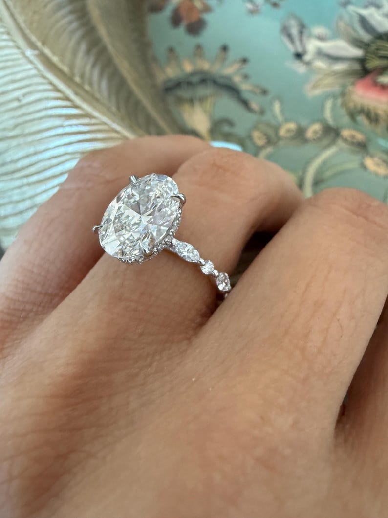 May include: A sparkling diamond ring with an oval-cut center stone. The ring features a delicate band with smaller accent diamonds. The setting is a bright, polished metal, likely platinum or white gold, and the ring is shown on a hand.