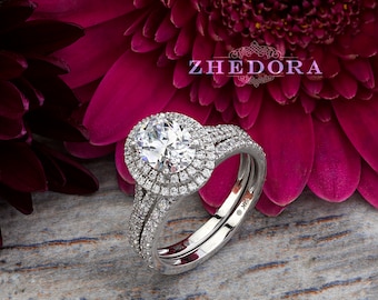 May include: A silver oval engagement ring with a double halo of smaller diamonds. The ring is set on a white background with pink flowers.