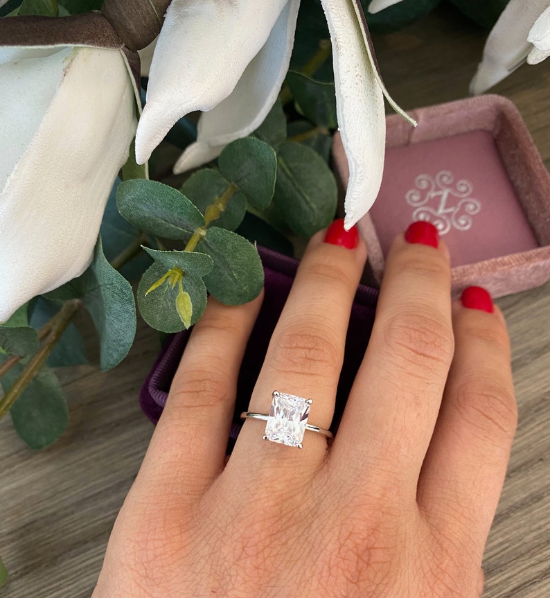 May include: A silver ring with a large, clear, rectangular-shaped diamond. The ring is on a finger with red nail polish.