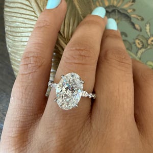 May include: A sparkling engagement ring featuring a large oval-cut diamond as the centerpiece. The ring has a delicate band with smaller accent stones, creating a brilliant display of light. The ring is set on a hand with light blue nail polish.
