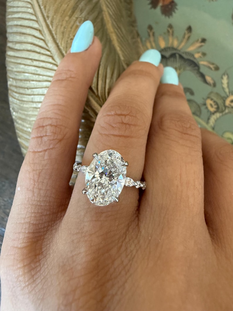 May include: A sparkling engagement ring featuring a large oval-cut diamond as the centerpiece. The ring has a delicate band with smaller accent stones, creating a brilliant display of light. The ring is set on a hand with light blue nail polish.