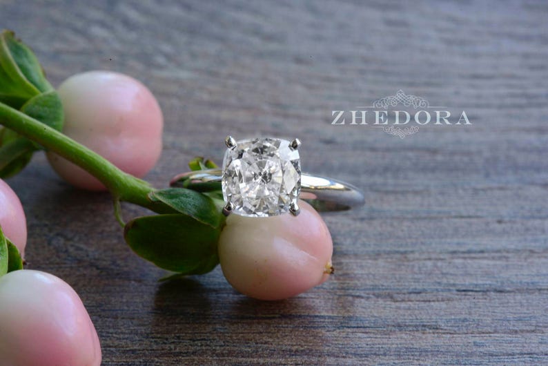 May include: A silver ring with a large, clear, oval-shaped diamond. The ring is set on a wooden surface with pink flowers.