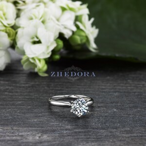 May include: A silver solitaire engagement ring with a round brilliant cut diamond set in a six-prong setting. The ring is on a gray wooden surface.