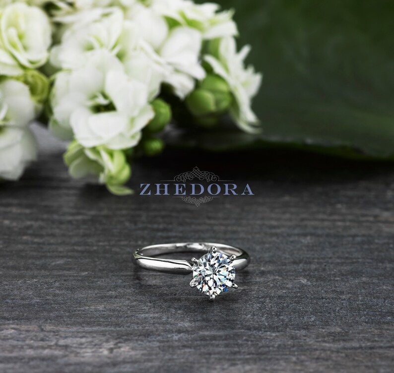 May include: A silver solitaire engagement ring with a round brilliant cut diamond set in a six-prong setting. The ring is on a gray wooden surface.
