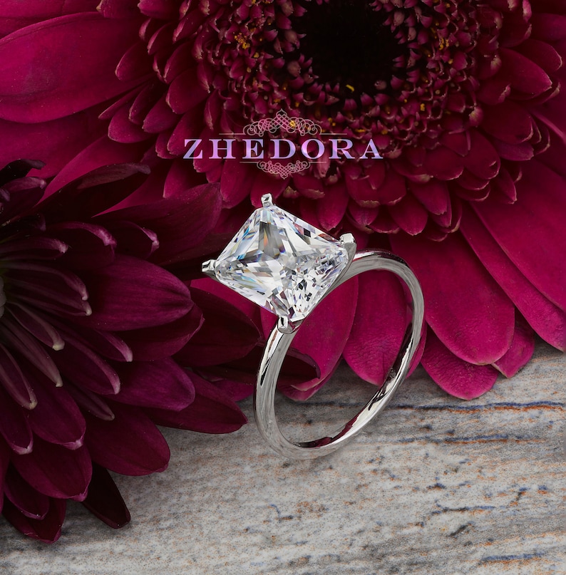 May include: A silver ring with a square-shaped, clear, princess-cut diamond. The ring is set against a background of pink flowers.