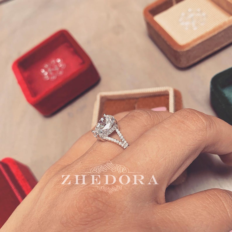 May include: A silver engagement ring with a large oval-shaped diamond and smaller diamonds set in the band. The ring is on a finger and the word "ZHEDORA" is visible in the background.