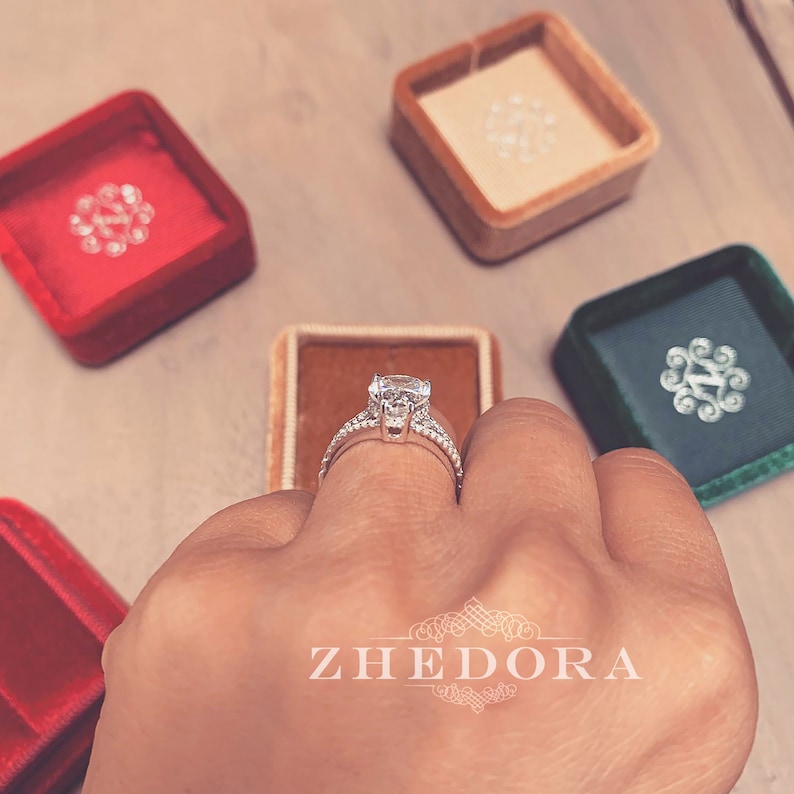 May include: A close-up of a silver engagement ring with a large, round, clear diamond. The ring is on a finger and there are several velvet ring boxes in the background.