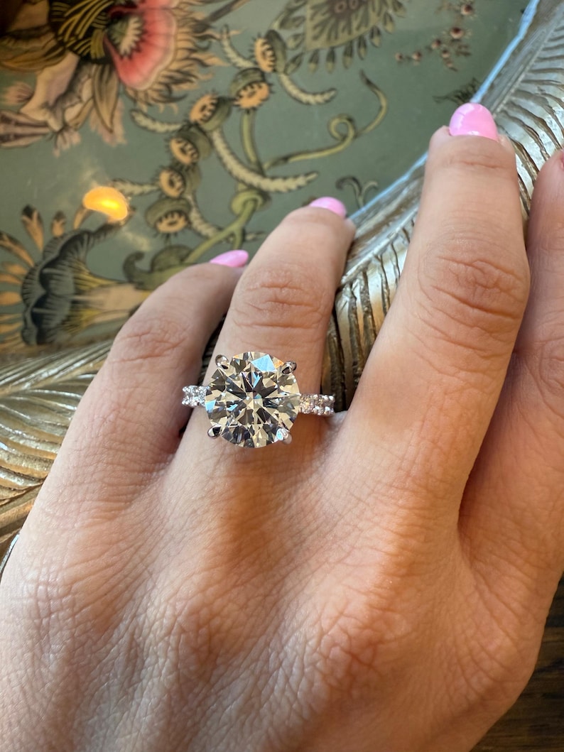 K&ouml;nnte beinhalten: Ein Platinring mit einem gro&szlig;en, runden, brillantgeschliffenen Diamanten als Mittelpunkt. Der Ring hat kleinere Akzentdiamanten auf beiden Seiten des Hauptsteins. Der Ring befindet sich an einer Hand mit rosa Nagellack.