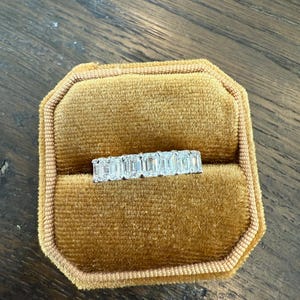 May include: A diamond eternity band ring with emerald cut stones set in a gold band. The ring is displayed in a brown velvet ring box.