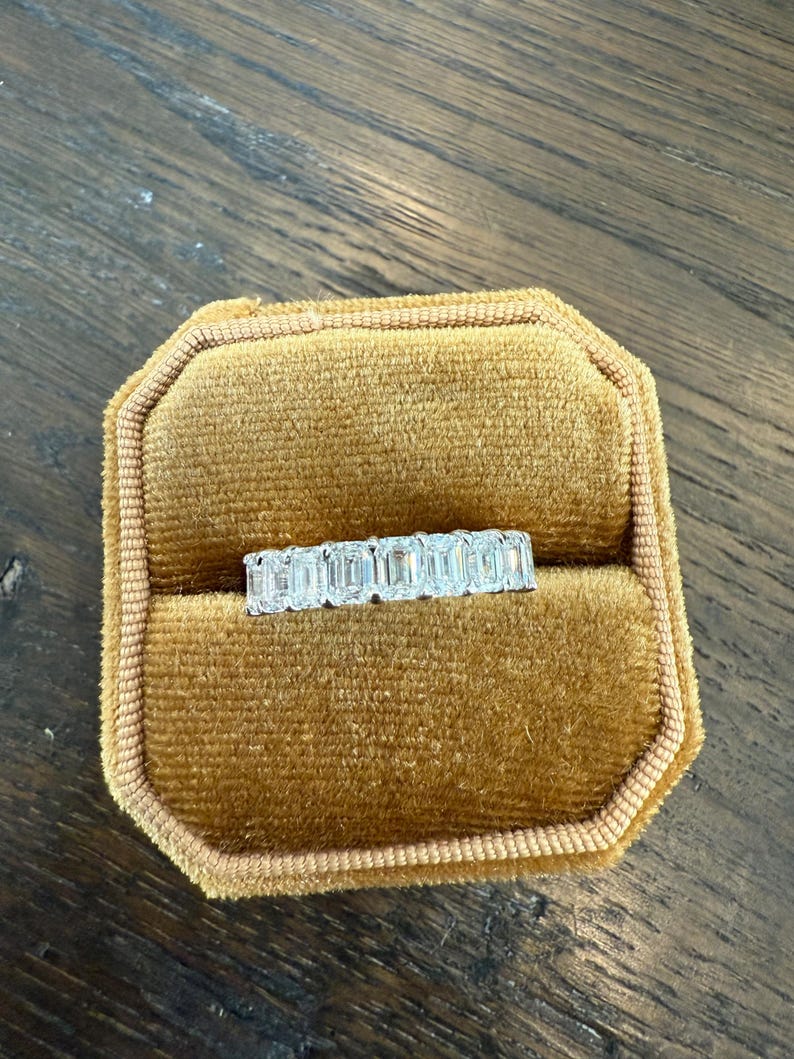 May include: A diamond eternity band ring with emerald cut stones set in a gold band. The ring is displayed in a brown velvet ring box.
