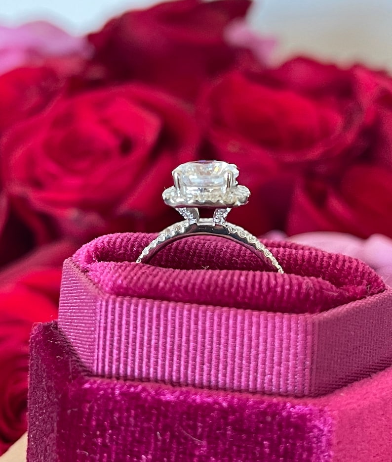 May include: A silver engagement ring with a large, clear, round diamond center stone surrounded by a halo of smaller diamonds. The ring is sitting in a pink velvet ring box.