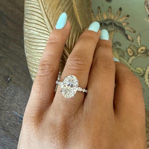 May include: A diamond engagement ring with an oval-cut center stone and a delicate band. The ring is set on a hand with light blue nail polish. The ring's design features smaller accent stones along the band.