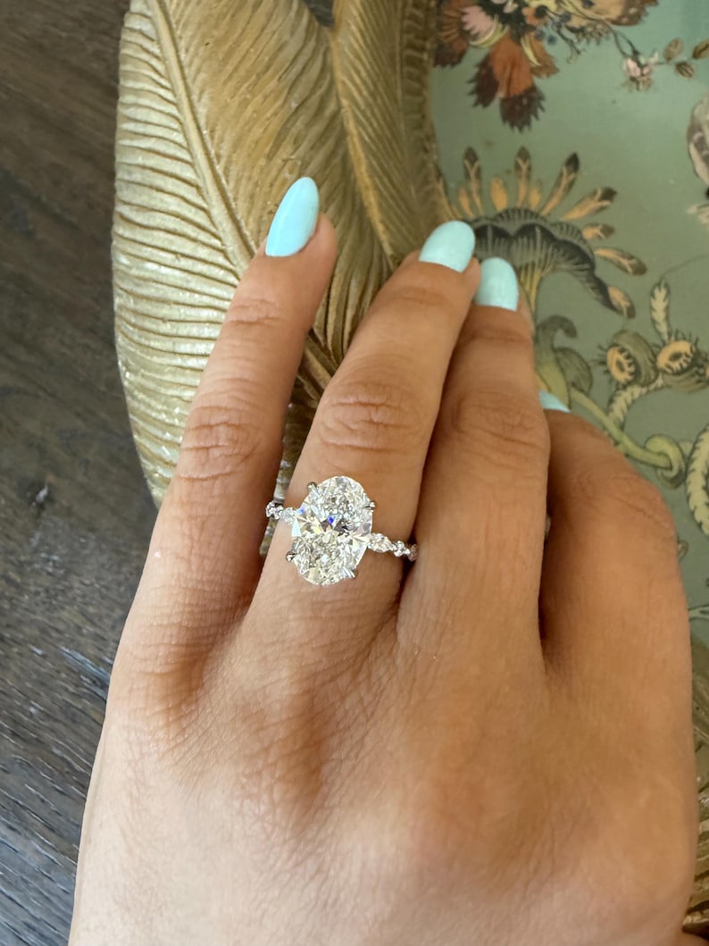 May include: A diamond engagement ring with an oval-cut center stone and a delicate band. The ring is set on a hand with light blue nail polish. The ring's design features smaller accent stones along the band.