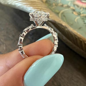 May include: A close-up of a silver engagement ring with a large, round, clear gemstone as the centerpiece. The ring features a halo of smaller stones around the main stone and additional stones set into the band.