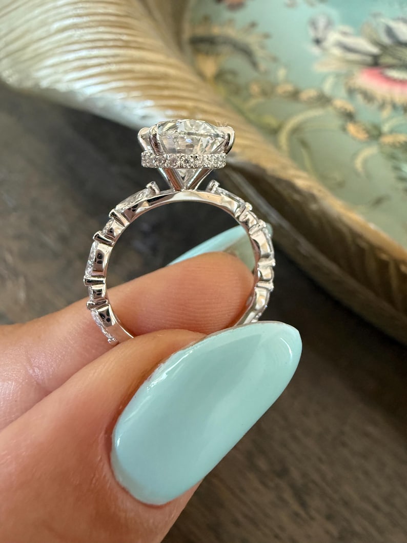 May include: A close-up of a silver engagement ring with a large, round, clear gemstone as the centerpiece. The ring features a halo of smaller stones around the main stone and additional stones set into the band.
