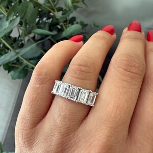 May include: A silver band ring featuring a row of emerald-cut diamonds. The ring is worn on a person's finger.