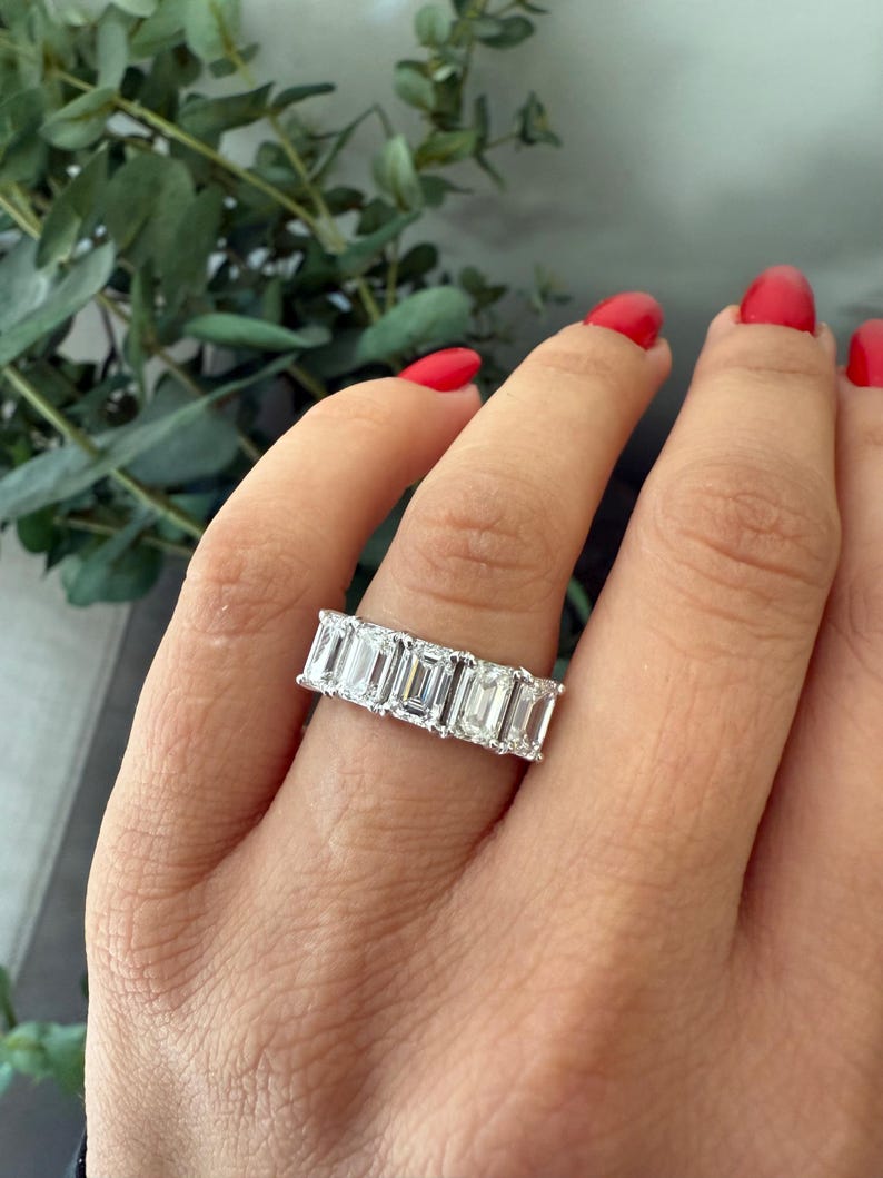 May include: A silver band ring featuring a row of emerald-cut diamonds. The ring is worn on a person's finger.