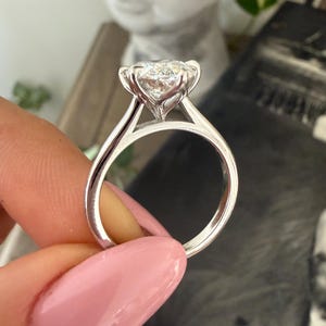 Pu&ograve; includere: Primo piano di un anello di fidanzamento in argento con un grande diamante rotondo e trasparente. L'anello ha una delicata montatura a griffe e una fascia liscia e lucida. L'anello &egrave; tenuto da una persona con lo smalto rosa.
