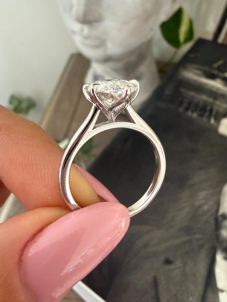 Pu&ograve; includere: Primo piano di un anello di fidanzamento in argento con un grande diamante rotondo e trasparente. L'anello ha una delicata montatura a griffe e una fascia liscia e lucida. L'anello &egrave; tenuto da una persona con lo smalto rosa.