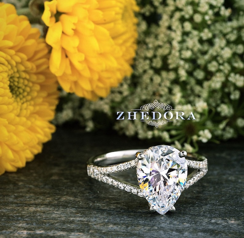 May include: A pear-shaped diamond engagement ring with a split shank and pave diamonds. The ring is set in white gold and features a large, sparkling diamond.
