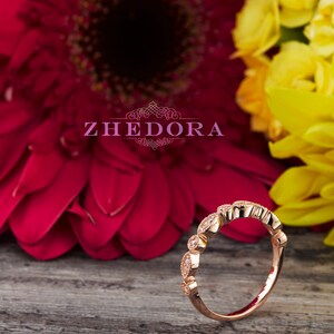 May include: A delicate rose gold band ring with a row of small, clear gemstones set in a scalloped design. The ring is photographed on a wooden surface with pink and yellow flowers in the background.