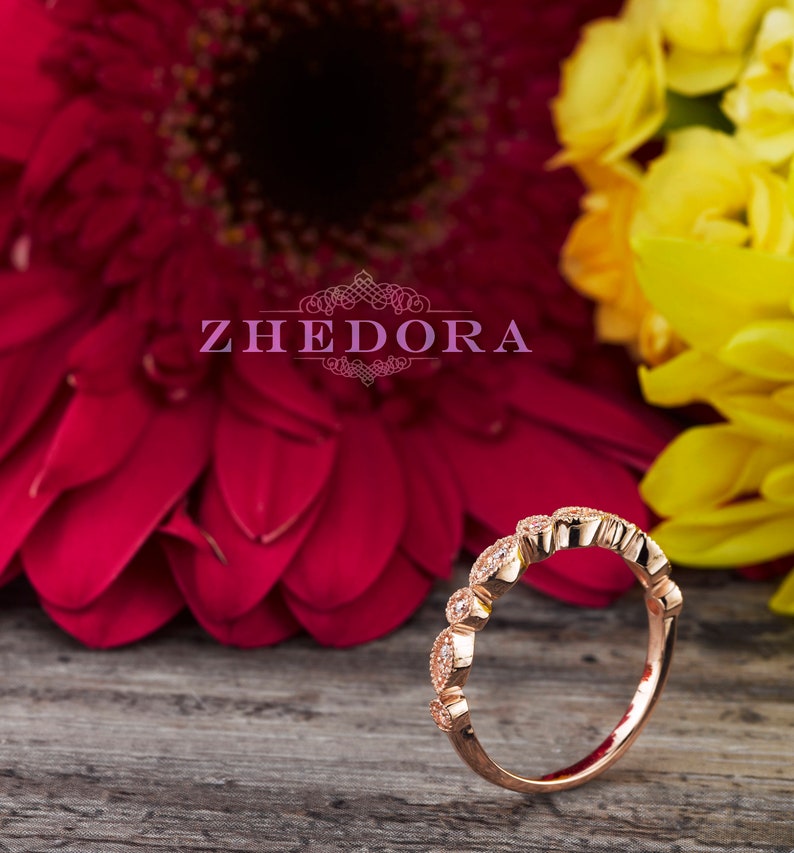 May include: A delicate rose gold band ring with a row of small, clear gemstones set in a scalloped design. The ring is photographed on a wooden surface with pink and yellow flowers in the background.