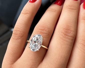 Könnte beinhalten: Ein goldener Ring mit einem großen, oval geschliffenen, klaren Edelstein, der von vier zarten Krappen gehalten wird. Die Ringschiene ist dünn, poliert und rund und zeigt ein klassisches Design, das den Glanz des Steins betont.