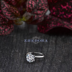 May include: A silver solitaire engagement ring with a large, round, clear diamond. The ring is set on a simple band and is photographed on a dark, textured surface.