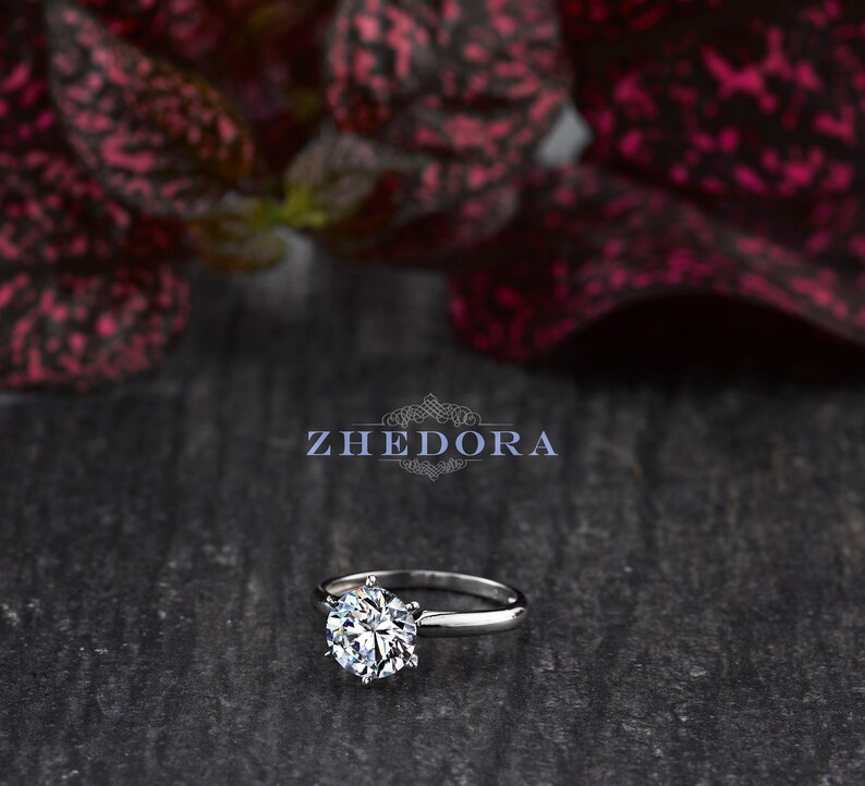 May include: A silver solitaire engagement ring with a large, round, clear diamond. The ring is set on a simple band and is photographed on a dark, textured surface.