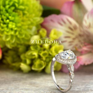 May include: A pear-shaped diamond engagement ring with a halo setting and a delicate, twisted band. The ring is set in silver and features a row of small diamonds on the band.